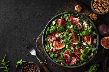 Salad with figs, prosciutto ham, arugula, goat cheese, nuts and pomegranate seeds with fork on black background. top view