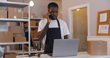 Mixed-race man talking on phone using laptop working at warehouse for online seller