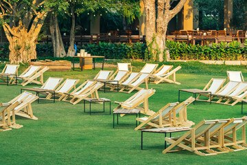 Many empty white deck chairs with tables in lawn is surrounded by shady green grass. Comfortable on...