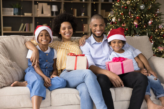 Happy young loving african american couple parents cuddling small children, sitting together on couch with wrapped Christmas gifts in hands near decorated tree at home, winter holidays concept.