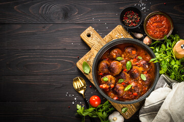 Meatballs in tomato sauce in a skillet on dark wooden table with ingredients. Top view with copy space.