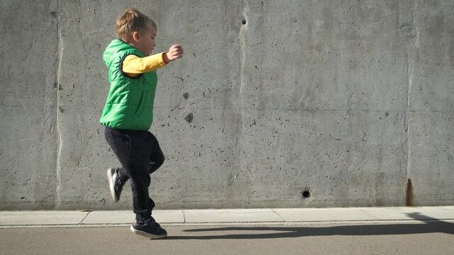 Cool modern preschool child boy in yellow and green clothes dancing near grey concrete wall in the city in sunny day. Funny ecstatic kid jumping and having fun in slow motion.