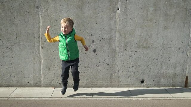 Cool trendy modern preschool child boy in yellow and green clothes dancing near grey concrete wall in the city in sunny day. Funny ecstatic kid jumping and having fun in slow motion.