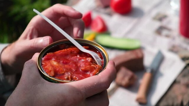 A man puts a fork in a jar of canned fish in tomato sauce. In the background is an impromptu table with vegetables and bread. Fast food, delicious snacks. Picnic in nature.