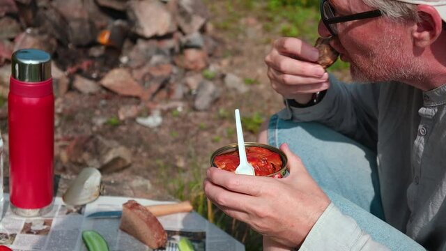 Picnic in nature. A middle-aged man with glasses is happy to eat canned fish in tomato sauce and bread. In the background is an impromptu table with vegetables and bread. Fast food, delicious snacks.