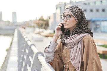 Young elegant woman in muslim clothing having a conversation on mobile phone while standing in the...