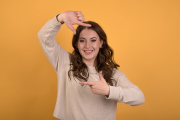 A young and attractive Caucasian brunette girl with wavy hair in a casual jumper making a photo frame with her hands on an orange studio background.