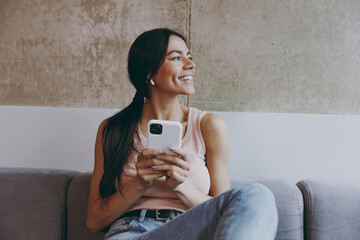 Happy bright young latin woman 20s in casual clothes sit on sofa listen music in earphones hold use mobile cell phone look aside spend time in living room home own house be lost in reverie good day