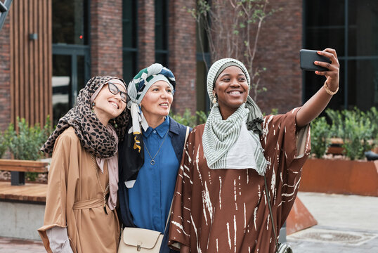 Three elegant women in muslim clothes smiling at camera of mobile phone while making selfie portrait outdoors