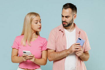 Young couple two friends family sad man jelaous woman wearing casual clothes peeps hold in hand use mobile cell phone together isolated on pastel plain light blue color background studio portrait