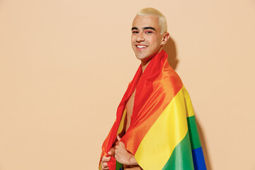 Side view young smiling happy blond latin gay man with make up in beige tank shirt wrapped in rainbow flag looking camera isolated on plain light ocher background studio People lgbt lifestyle concept.