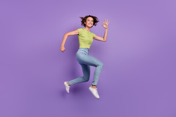 Fototapeta na wymiar Photo of impressed sweet young lady wear green t-shirt showing fingers okey jumping high isolated violet color background