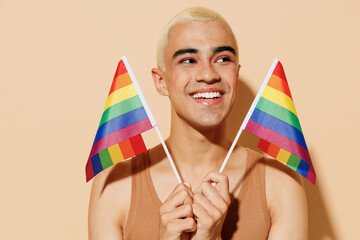 Young minded happy blond latin gay man with make up in beige tank shirt holding rainbow flag look aside isolated on plain light ocher color background studio portrait People lgbt lifestyle concept.