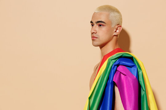 Side view young minded blond latin gay man 20s with make up in beige tank shirt with rainbow flag on shoulder look aside isolated on plain light ocher background studio People lgbt lifestyle concept.