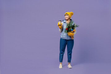 Full size body length mature elderly woman 55 years old wears blue waistcoat yellow hat mittens holds spruce branches craft paper cup isolated on plain pastel light violet background studio portrait.