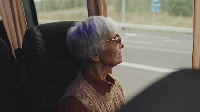 Medium close-up of senior gray-haired woman in dark sunglasses sitting by window during bus ride