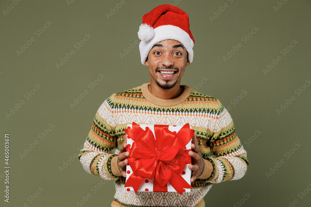 Wall mural young african man in knitted sweater red christmas santa claus hat hold red present box with gift ri