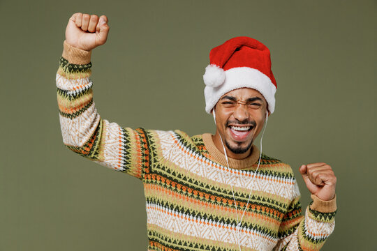 Young african man in knitted sweater red Christmas Santa Claus hat headphones listen to music dance have fun isolated on plain green khaki background studio. Happy New Year 2022 celebration concept.