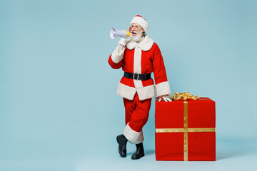 Full body old Santa Claus man 50s wearing Christmas hat red suit clothes stand near gift box scream in megaphone isolated on plain blue background studio. Happy New Year 2022 merry ho x-mas concept.