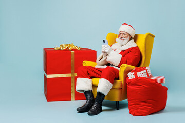 Full size old Santa Claus man in Christmas hat red suit clothes sit in chair use mobile cell phone isolated on plain blue background studio. Happy New Year celebration merry ho x-mas holiday concept.