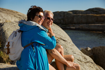 Loving mature couple traveling, sitting on the rock, exploring. Real man and woman hugging, kissing, Happily smiling. Scenic view of Serene Scandinavian landscape on south coast of Norway, Tjøme.