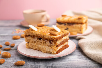 Honey cake with milk cream, caramel, almonds and a cup of coffee on a gray and pink background. Side view, selective focus.