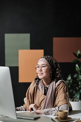 Young muslim businesswoman sitting at the table looking at computer monitor and typing, she working...