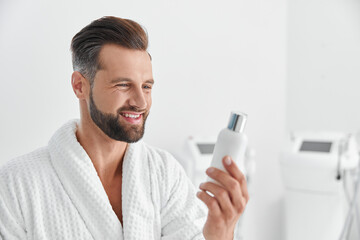 Happy man in white gown looks at bottle of cosmetic product in cosmetologycal clinic