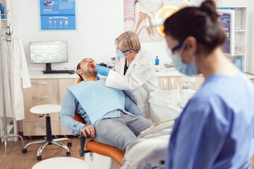 Senior stomatologist holding sterilized dental tools talking with sick patient before stomatological intervation. Nurse and doctor wearing masks cleaning teeth of man sitting on chair with open mouth