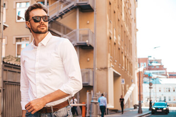 Portrait of handsome confident stylish hipster lambersexual model.Modern man dressed in white shirt. Fashion thoughtful male posing on the street background. Outdoors at sunset in sunglasses