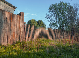 Old wooden fence in the garden plot against the background of green grass, old wooden house and blue sky