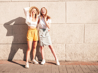 Two young beautiful smiling hipster female in trendy summer clothes.Sexy carefree women posing near white wall in the street. Positive pure models having fun at sunset, hugging and going crazy