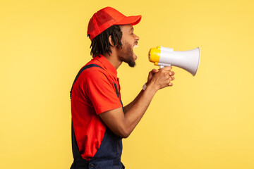 Side view portrait of aggressive worker wearing red T-shirt, cap and blue overalls holding megaphone and screaming loud, protesting. Indoor studio shot isolated on yellow background.