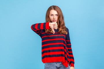 Unhappy unsatisfied young woman wearing striped casual style sweater showing thumbs down, dislike...