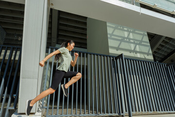 Graceful sportive Asian woman in stylish tracksuit jumping upward moment at training near modern multi-storey parking fence on sunny day