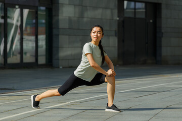 Positive strong Asian woman in tracksuit and shoes does forward dynamic lunges training on sunny...
