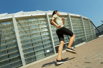 Fototapeta na wymiar Slim young Asian woman in comfortable tracksuit runs along road past modern stadium building under clear sky on sunny day
