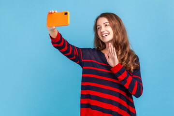 Portrait of cheerful woman wearing striped casual style sweater, broadcasting livestream, waving hand, saying hi to followers, or having video call. Indoor studio shot isolated on blue background.