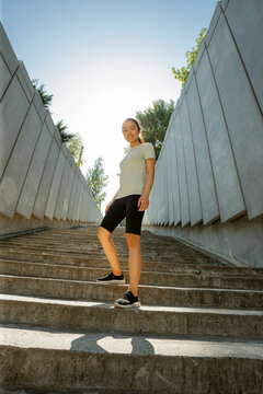 Confident Young Asian Woman In Stylish Tracksuit Stands On Stone Staircase After Training At Sunrise On Street Low Angle Shot