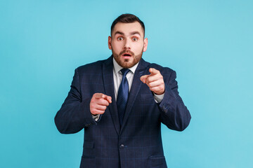 Amazed astonished bearded man wearing official style suit pointing finger to camera, looking with interrogative expression at camera. Indoor studio shot isolated on blue background.