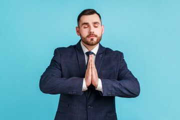 Portrait of calm relaxed young man with beard wearing official style suit standing with raised arms and doing yoga meditating exercise. Indoor studio shot isolated on blue background.