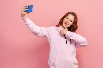 Portrait of smiling curly-haired teen girl in hoodie taking selfie, having conversation on video call or streaming blog, pointing finger. Indoor studio shot isolated on pink background