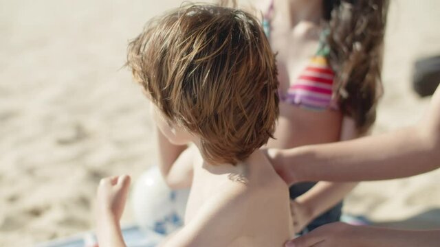 Slow Motion Of Elder Sisters Hands Putting Sunblock On Boys Back. Sad And Pensive Brother Upset That Sisters Applying Sunscreen Lotion On His Skin During Summer Vacation. Skincare, Family Concept