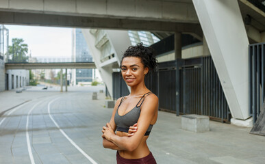 Cheerful biracial lady in stylish top with crossed arms stands against contemporary city building fence
