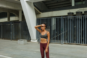 Tired biracial lady with bare tummy in stylish top touches forehead resting at training against contemporary city building fence
