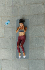 Brunette woman in stylish tracksuit does abdominal exercises on mat at training on street pavement view from above