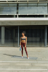 Fototapeta na wymiar Young biracial sportswoman in stylish top stands resting during training on empty road against building metal gate