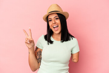 Young caucasian woman with one arm wearing a summer hat isolated on pink background joyful and carefree showing a peace symbol with fingers.