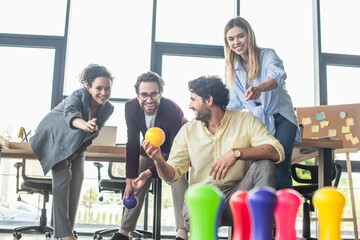 Businessman holding bowling ball near smiling interracial colleagues in office