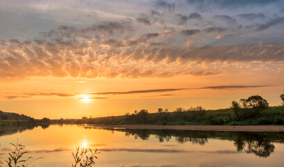 Naklejka na ściany i meble Scenic view at beautiful summer river sunset with reflection on water with green bushes, calm water ,deep colorful cloudy sky and glow on horizon on a background, spring evening landscape